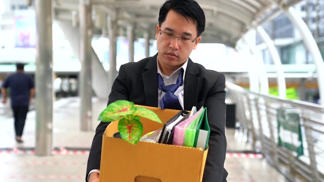 Unhappy businessman walking and carrying his belongings in a paper box after being fired.