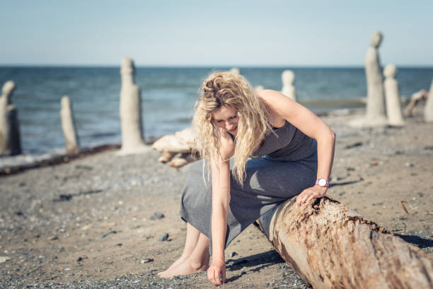 A young woman picking up a pebble from the seashore, sitting on a tree trunk on a sunny day