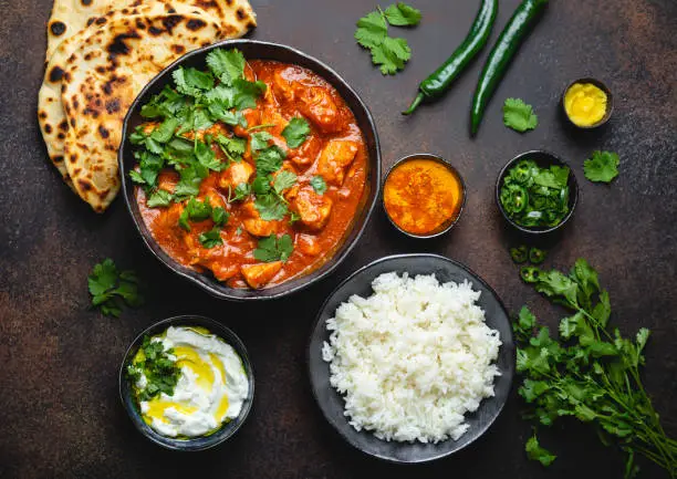 Traditional Indian dish Chicken tikka masala with spicy curry meat in bowl, basmati rice, bread naan, yoghurt raita sauce on rustic dark background, top view, close up. Indian style dinner from above