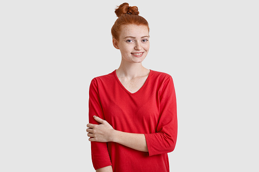 Indoor shot of glad red haired woman with hair bun, keeps hands partly crossed, wears red sweater, has freckled skin, enjoys pleasant conversation, models over white background. Good emotions