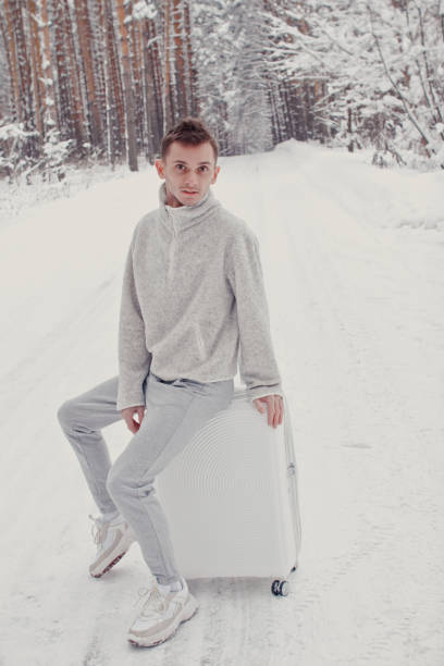 Portrait of young fashionable man on snow holding suitcase and showing thumbs up. Winter vacation travel concept. Portrait of young fashionable man in white clothes walking on snow holding suitcase and go pathway pine forest. Winter vacation travel concept. Outdoors in winter. Ski resort, mounting skiing resort thumbs up for skiing stock pictures, royalty-free photos & images