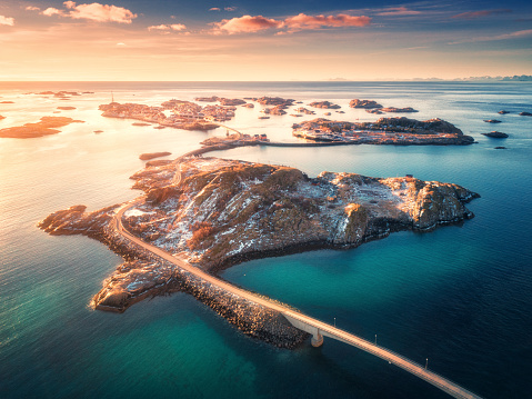Aerial view of bridge over the sea and mountains in Lofoten Islands, Norway. Henningsvaer at sunset in winter. landscape with azure water, sky with gold sunlight, rocks, buildings, road. Top view