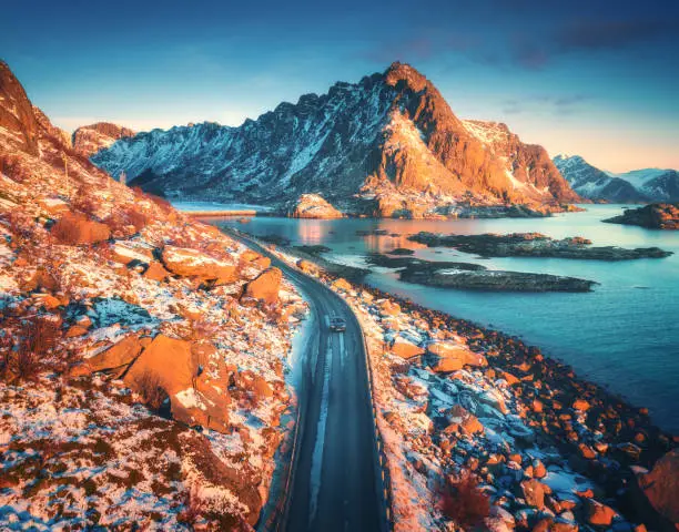 Aerial view of beautiful mountain road near the sea, mountains, purple sky at sunset in Lofoten islands, Norway in winter. Top view of road, car, high snowy rocks with stones, coastline, blue water