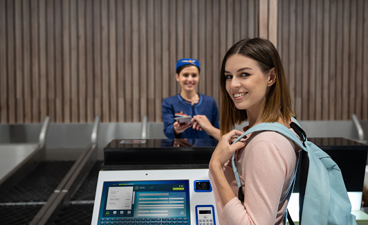 Portrait of a happy female traveler doing the check-in at the airport getting her boarding pass at the counter and looking at the camera smiling - travel concepts. **DESIGN ON SCREEN WAS MADE FROM SCRATCH BY US**