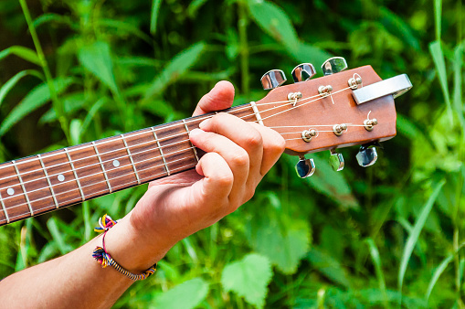 Mans hand holding chord E on a wooden acoustic guitar neck fingerboard fretboard on scenic green flora background