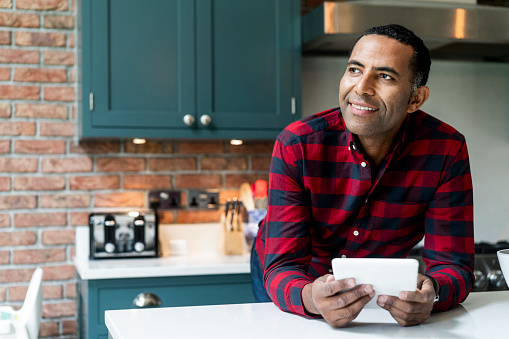 Thoughtful mature man leaning on kitchen island. Smiling male is holding digital tablet while standing in kitchen. He is in red checked shirt at home.