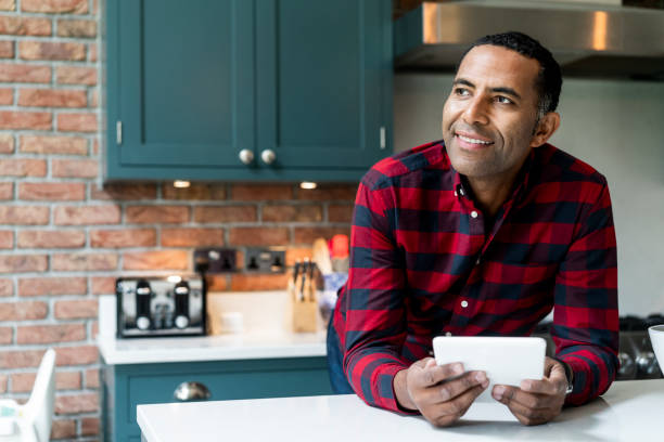 Thoughtful man with digital tablet in kitchen Thoughtful mature man leaning on kitchen island. Smiling male is holding digital tablet while standing in kitchen. He is in red checked shirt at home. 45 49 years stock pictures, royalty-free photos & images