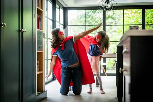 Playful mother and daughter in superhero costume against window. Woman is kneeling with arms outstretched by girl standing in kitchen. They are enjoying at home.