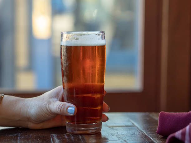 woman s hand with pink fingernails gripping an amber beer glass in front of window - amber beer imagens e fotografias de stock