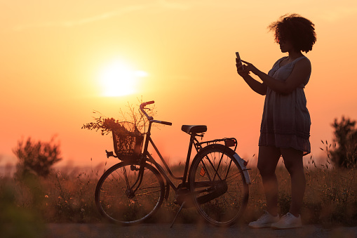 Silhouette of beautiful african woman with bike using smart phone, in nature.