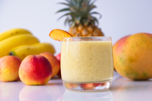 Healthy fruit smoothie in glass in front of fresh pineapple, bananas, peaches, mangoes.