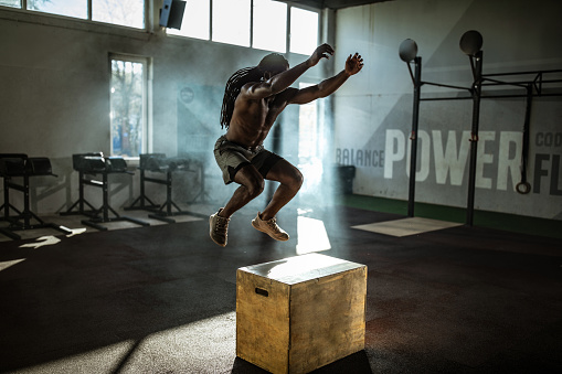 Black athletic man jumping on a crate while having sports training in a gym.