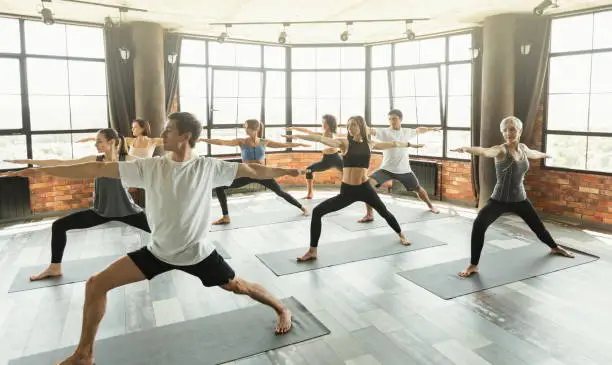 Group of young sporty people practicing yoga, standing in Warrior two pose, Virabhadrasana II pose, yogi millennials in a modern studio.