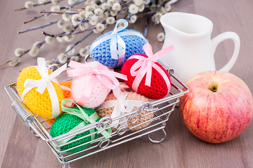 Knitted Easter eggs tied with colored ribbons in a metal basket, an apple, a jug and willow on a wooden table