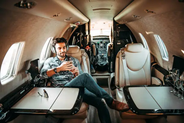 Young successful man listening to music over the headphones while sitting in a private airplane. Sunglasses are on the table in front of him.