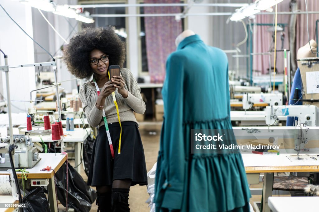 Female designer with tape measure using phone Female designer with tape measure working in fashion studio. African Ethnicity Stock Photo