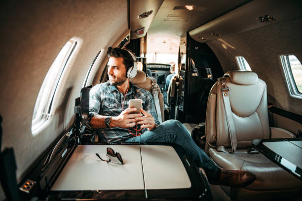 jeune homme riche qui regarde par la fenêtre tout en écoutant de la musique sur le casque et à l’aide d’un téléphone intelligent, assis dans un jet privé - status symbol audio photos et images de collection