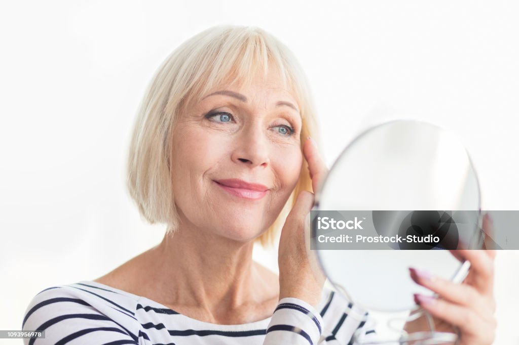Senior woman touching her soft face skin Senior woman touching her soft face skin, looking in mirror at home Aging Process Stock Photo
