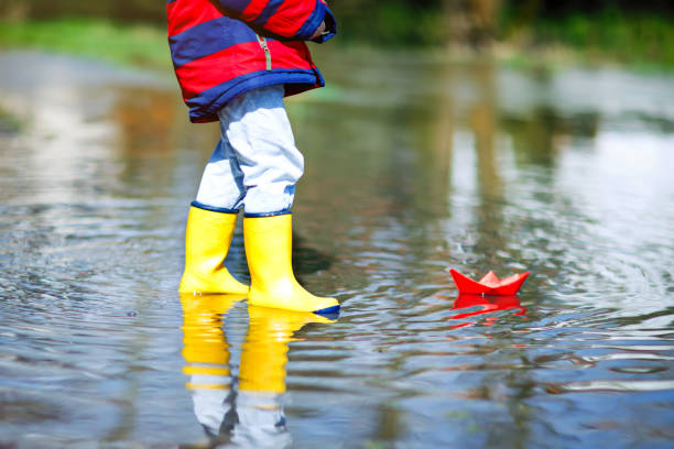 glückliche kleine kind junge in gelb regen stiefel mit papier schiff boot durch riesige pfütze auf frühjahr oder herbst tag spielen - puddle rain boot water stock-fotos und bilder