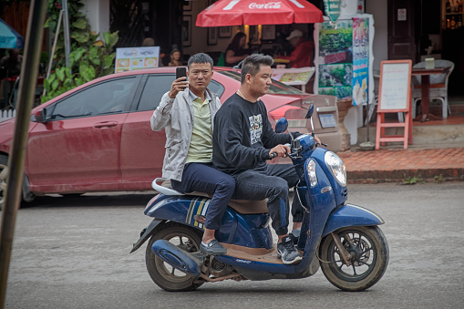 Sisavangvong Road, Luang Prabang, Laos - December 12, 2018: Couple of male tourists on a motorcycle  in main street of the former capital of Laos