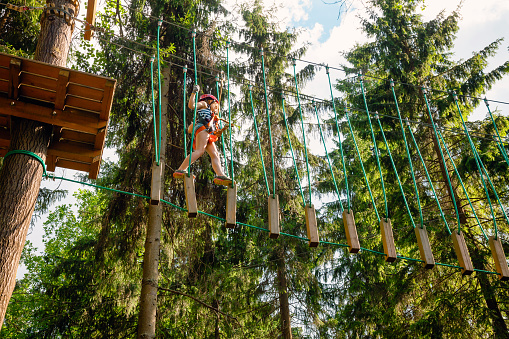Elementary age girl wearing safety harness passing unstable rope bridge obstacle at a ropes course in outdoor treetop adventure park