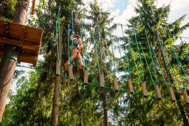 Little girl on a ropes course in a treetop adventure park passing hanging rope obstacle Elementary age girl wearing safety harness passing unstable rope bridge obstacle at a ropes course in outdoor treetop adventure park girl climbing ladder stock pictures, royalty-free photos & images