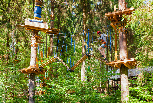 Teenager boy wearing safety harness passing rope bridge obstacle at a ropes course in outdoor treetop adventure park