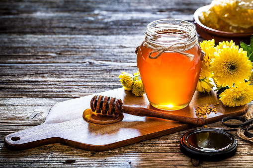 Apiculture industry concepts. Organic honey in a glass jar shot on rustic wooden table. The honey jar is on a small wooden cutting board and in front of it is a honey dipper with flowing honey. Yellow flowers are at the right and complete the composition. Useful copy space available for text and/or logo. Predominant colors are yellow and brown. Low key DSRL studio photo taken with Canon EOS 5D Mk II and Canon EF 100mm f/2.8L Macro IS USM.