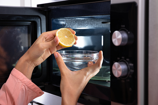 Woman's Hand Squeezing Halved Lemon In The Glass Bowl Near An Open Microwave Oven