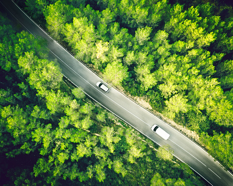 Aerial view from a drone of a two lane straight road in a beautiful green forest of pine trees in Europe and a white car and a van driving trough it. Horizontal image from RAW drone file in Spain. Sharp focus.