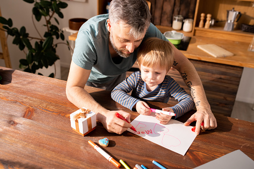 Serious concentrated bearded father and cute son standing at wooden table with markers and gift box and making Valentines card for mother