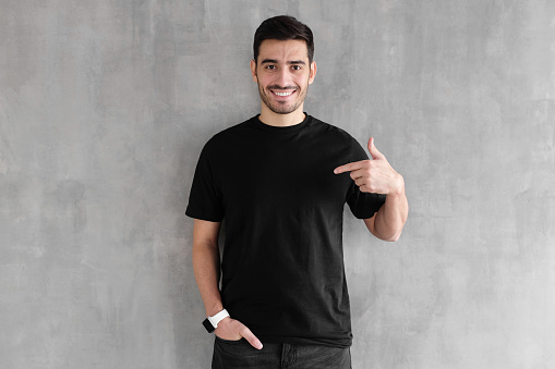 Young handsome man isolated on gray textured wall, smiling while pointing with index finger to black t-shirt, copyspace for advertising