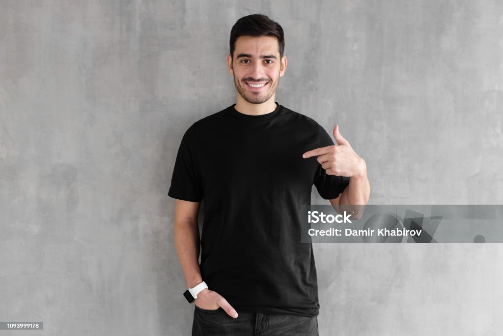 Young handsome man isolated on gray textured wall, smiling while pointing with index finger to black t-shirt, copyspace for advertising T-Shirt Stock Photo