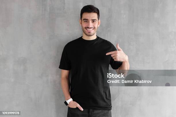 Young Handsome Man Isolated On Gray Textured Wall Smiling While Pointing With Index Finger To Black Tshirt Copyspace For Advertising Stock Photo - Download Image Now