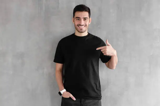 Photo of Young handsome man isolated on gray textured wall, smiling while pointing with index finger to black t-shirt, copyspace for advertising