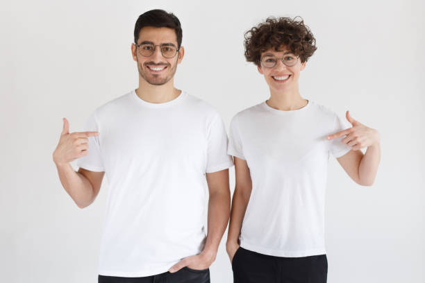 sonriente pareja joven apuntando a camisetas blancas en blanco con los dedos del índice, espacio de copia para su publicidad, aislado sobre fondo gris - de ascendencia europea fotografías e imágenes de stock
