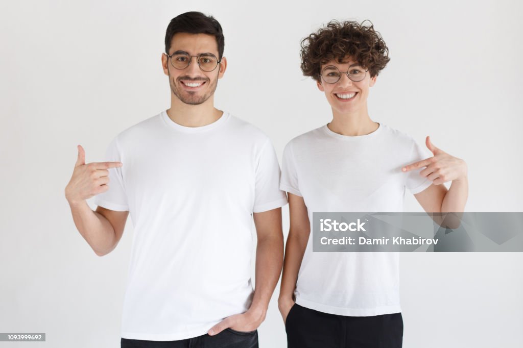 Souriante jeune couple pointant sur des t-shirts blancs blanc avec index, espace copie pour votre publicité, isolé sur fond gris - Photo de T-Shirt libre de droits