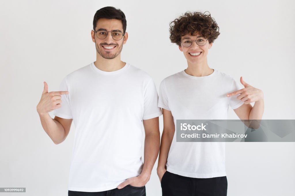 Smiling young couple pointing at blank white t-shirts with index fingers, copy space for your advertising, isolated on gray background T-Shirt Stock Photo