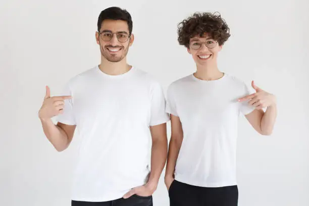 Photo of Smiling young couple pointing at blank white t-shirts with index fingers, copy space for your advertising, isolated on gray background