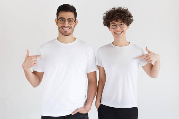 smiling young couple pointing at blank white t-shirts with index fingers, copy space for your advertising, isolated on gray background - white clothing imagens e fotografias de stock