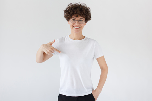 Smiling nice woman pointing at her blank white t-shirt with index finger, copy space for your advertising, isolated on gray background