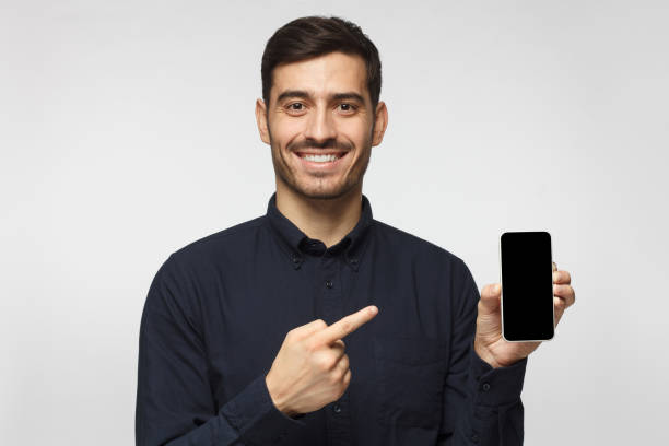 istock Handsome man in deep blue shirt isolated on gray background, presenting smartphone and pointing with finger at blank screen with copy space for ads 1093999794