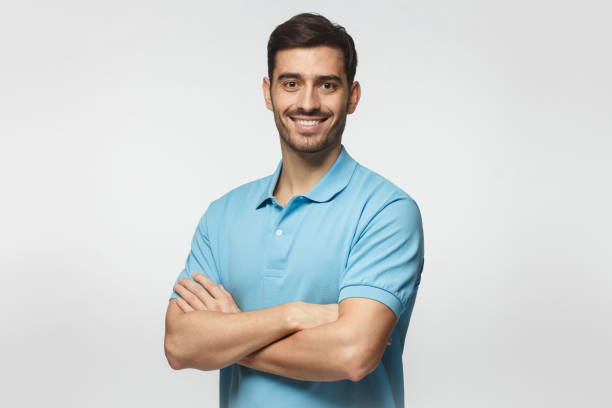 retrato de europeo caucásico joven aislado sobre fondo gris, pie en camisa polo azul con brazos cruzados, sonriendo y mirando a cámara - mens shirt fotografías e imágenes de stock