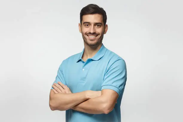 Photo of Portrait of young european caucasian man isolated on gray background, standing in blue polo shirt with crossed arms, smiling and  looking at camera