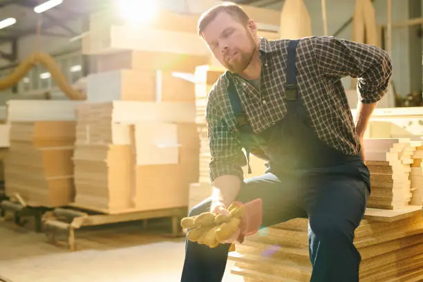 Tired handsome bearded manual worker with closed eyes sitting on stack of wooden plank and massaging lower back while feeling pain in back