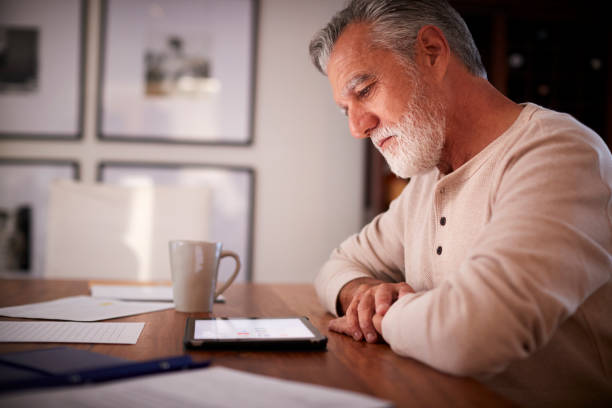 senior-hispanic mann sitzt an einem tisch mit einem tablet-computer am abend, nahaufnahme, seitenansicht - senior adult digital tablet domestic life learning stock-fotos und bilder