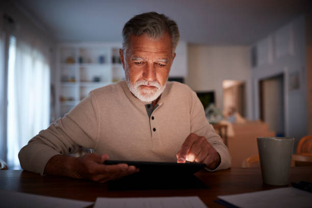 Senior Hispanic man sitting at a table reading an e book at home in the evening, close up