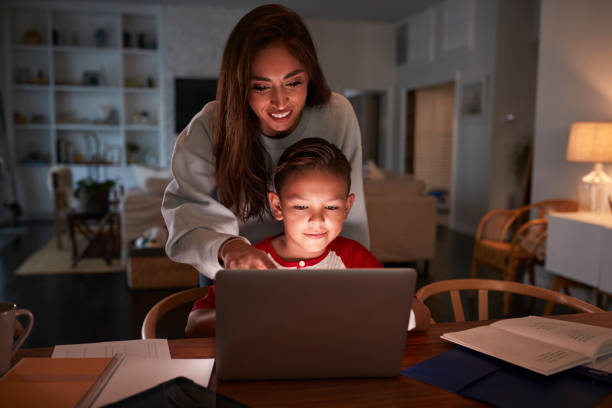 Hispanic woman looking over her sons shoulder while he does his homework using laptop computer Hispanic woman looking over her sonÕs shoulder while he does his homework using laptop computer low lighting stock pictures, royalty-free photos & images