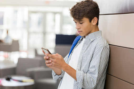 A serious teenage boy stands and leans against a wall in his high school lobby.  He texts a friend as he waits.