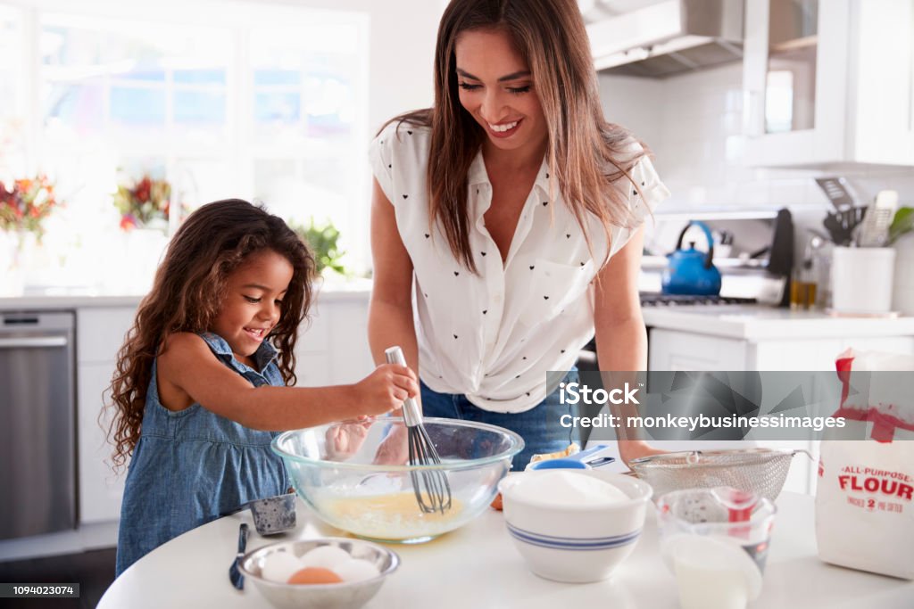 Young Hispanic girl making cake in the kitchen, overseen by her mum, waist up Child Stock Photo